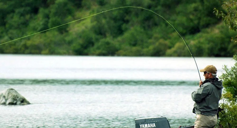 Angler with a bent fly rod, fighting a fish in a calm river.