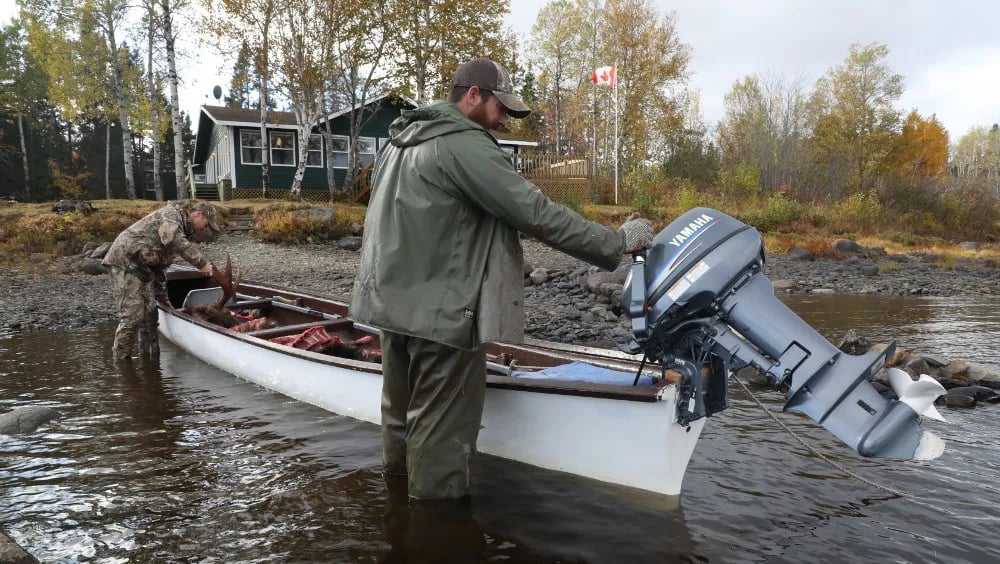 Two guides preparing a long boat in shallow river water with a rustic cabin and autumn trees in the background