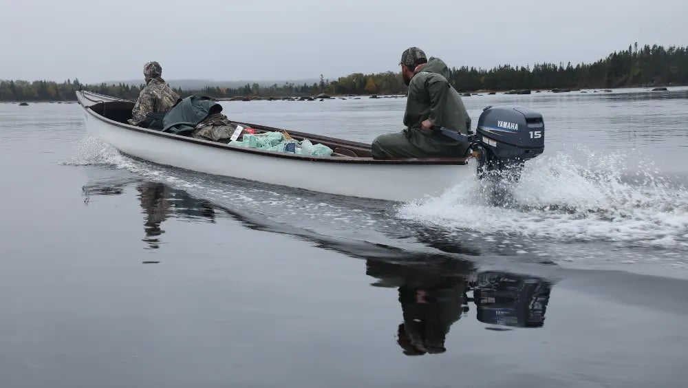Two men in a long boat speeding across a calm river, leaving a white wake behind them