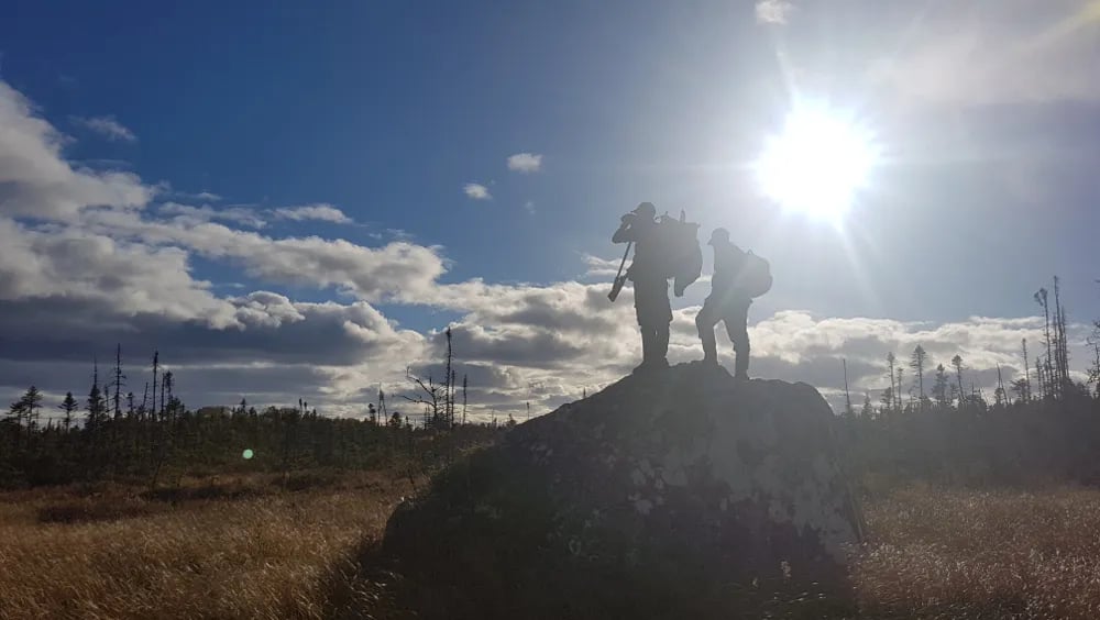Two people silhouetted on a grassy hill against a bright, cloudy sky with a strong sun flare