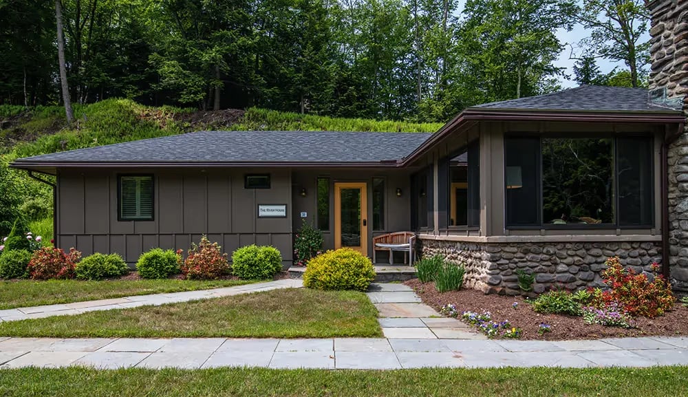 Modern single-story house with a stone foundation, brown vertical siding, a gray shingled roof, a bench near the entrance, and surrounding green landscaping.