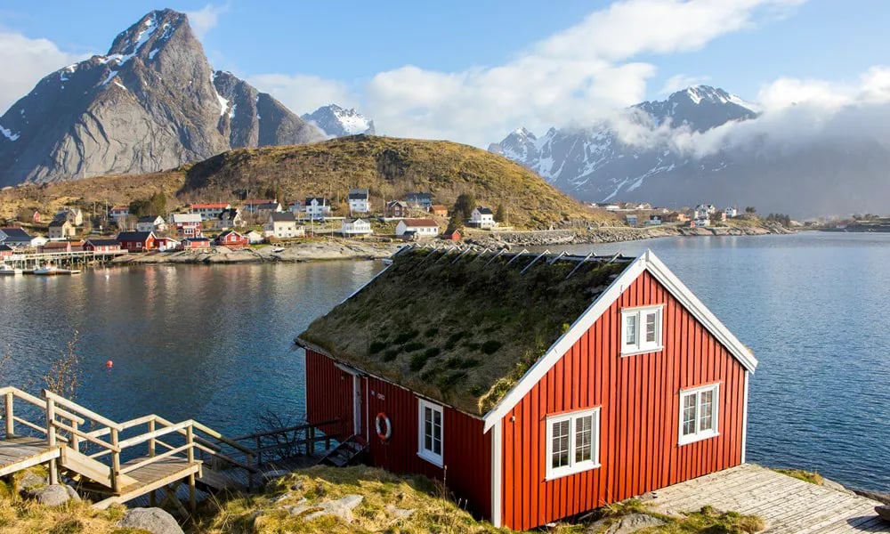 Red rorbu cabin with a grass roof on a point overlooking the fjord and mountains under a blue sky