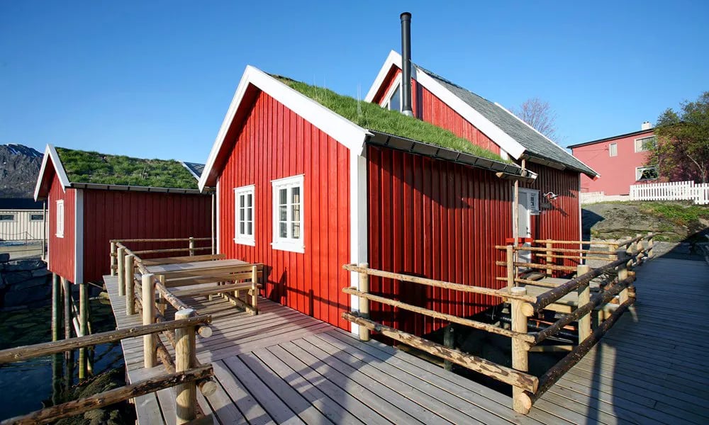 Red rorbu cabins with grass roofs connected by wooden boardwalks, set against a backdrop of mountains