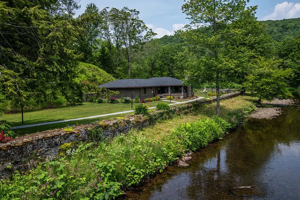 Single-story house with a dark roof surrounded by trees and greenery beside a clear stream with a stone wall in front.