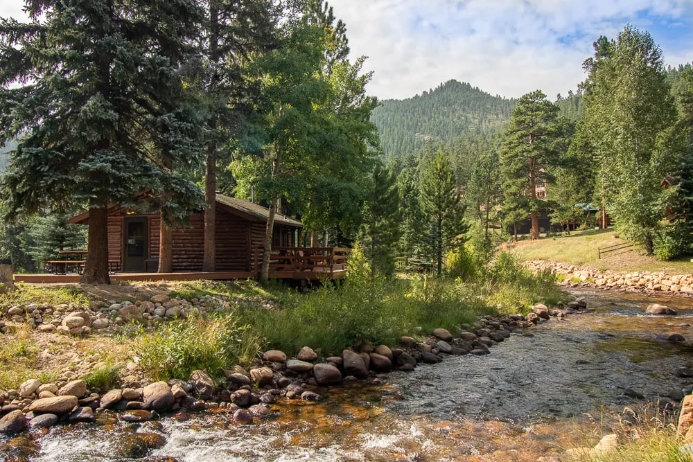 Rustic cabin exterior with a wooden deck next to a flowing river and pine trees