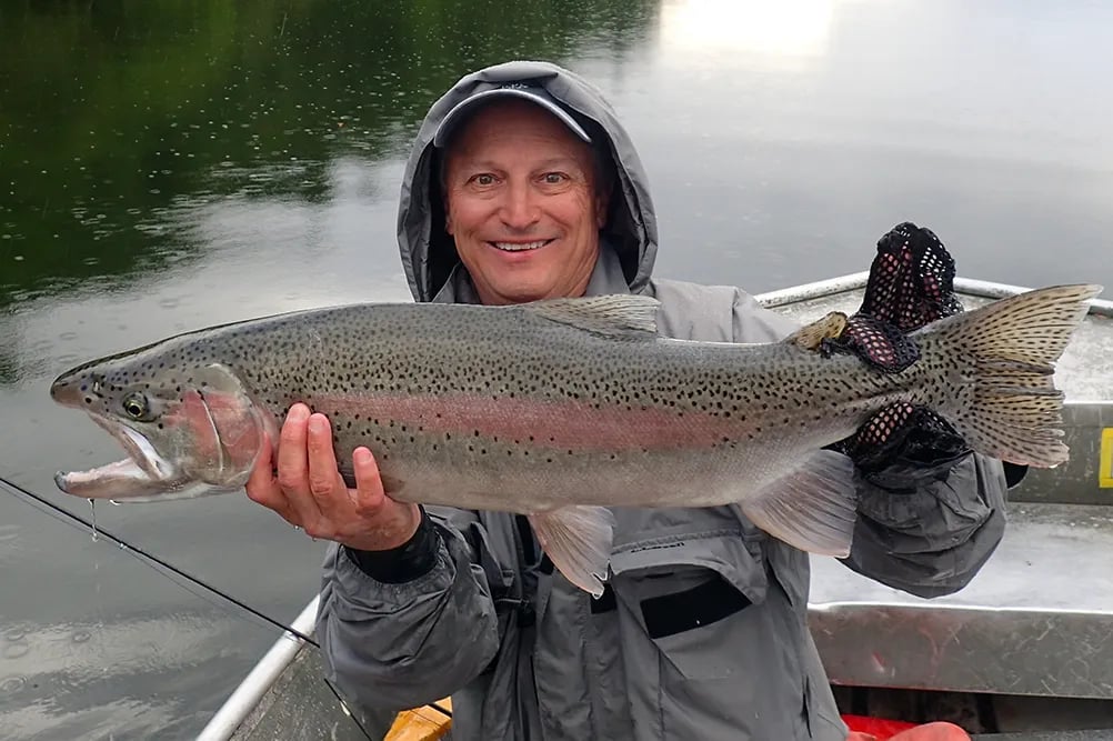 Smiling angler holding a large, iridescent rainbow trout in a boat.