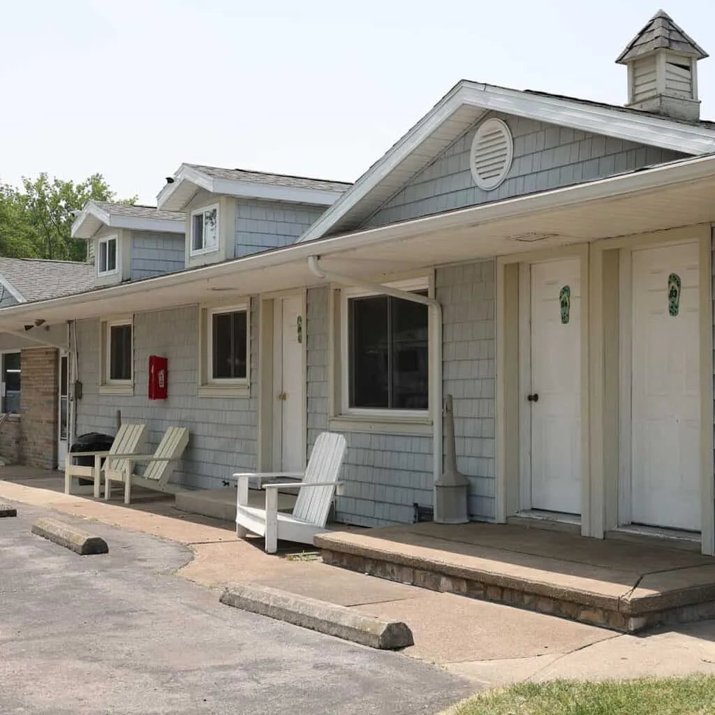 Exterior of a light-blue motel building with multiple rooms and white Adirondack chairs on the porch