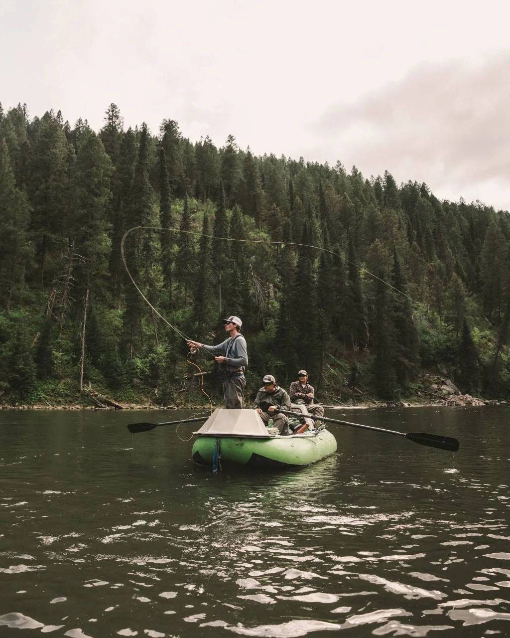 Angler casting a fly rod from a green drift boat on a tree-lined river