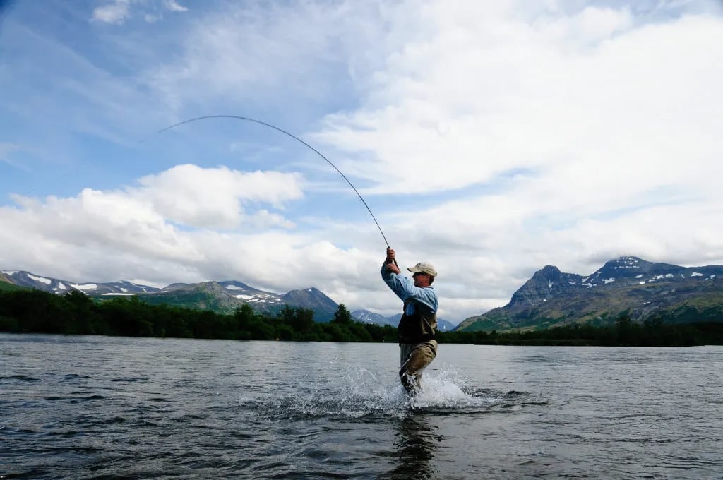 Angler in waders casting a fly rod in a wide river with a dramatic mountain range under a cloudy sky