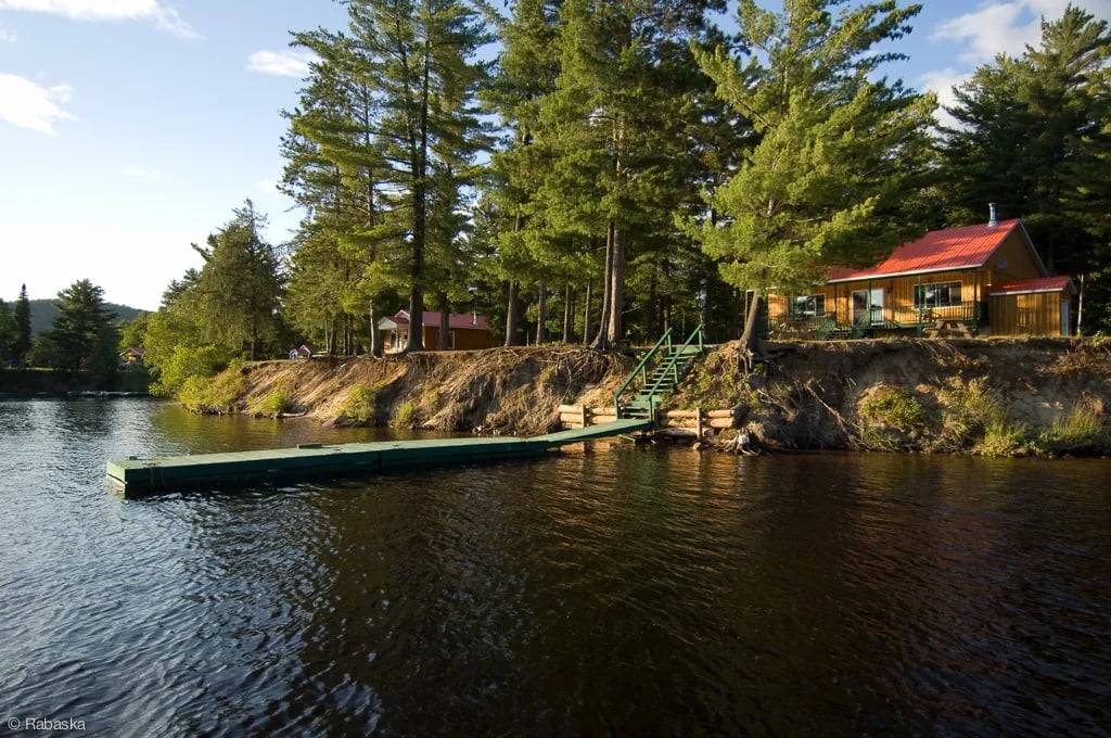 Wooden cottage on a high bank overlooking a calm river, with a floating dock.
