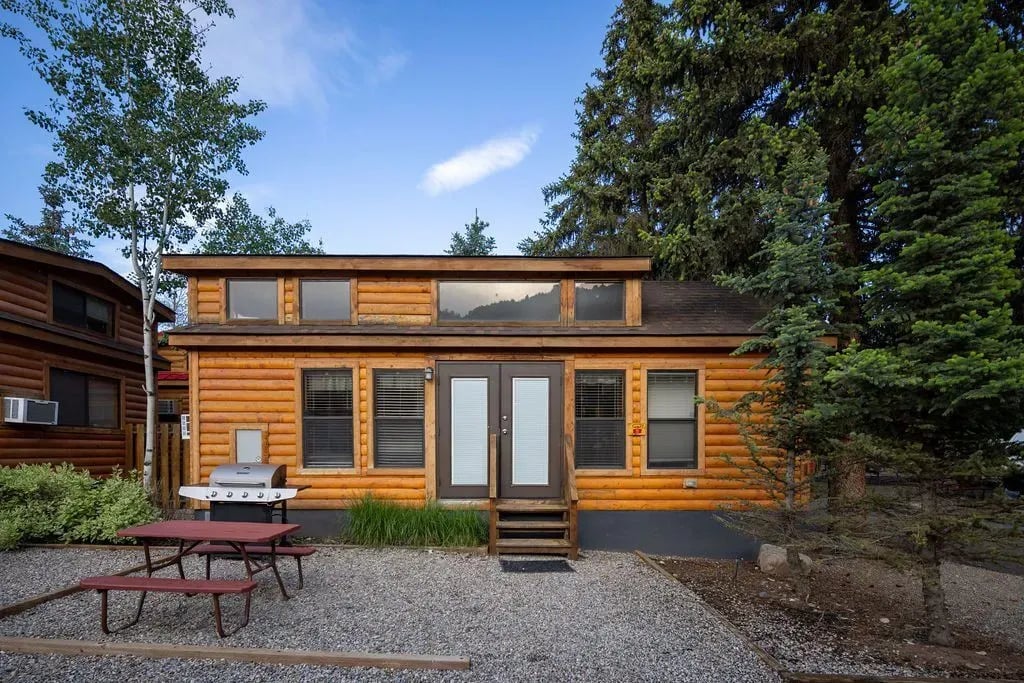 Log cabin exterior with a picnic table, grill, and tall trees.