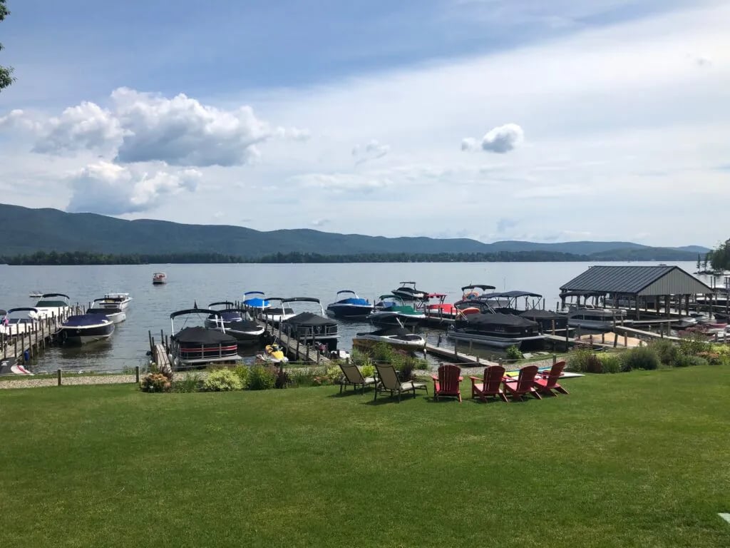 Lakeside marina with numerous boats docked, green lawn, and distant mountains