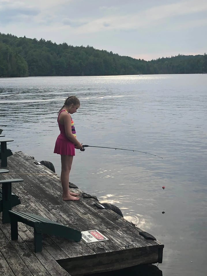 Girl in a kayak with a dog on a dock, calm lake in background.