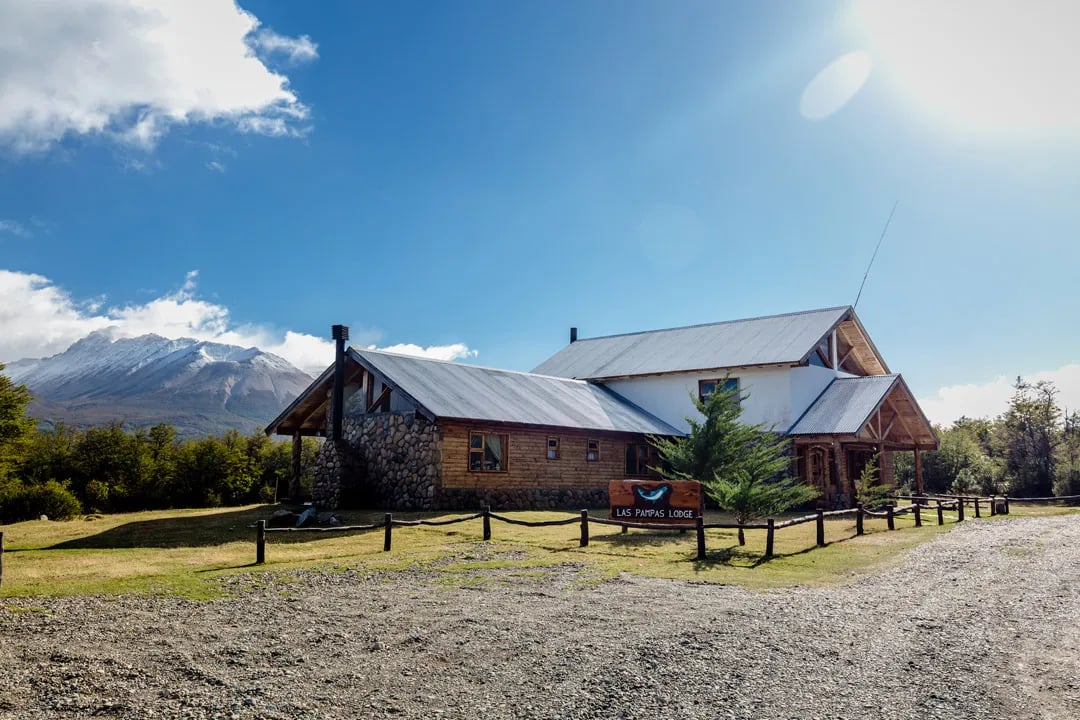 Rustic lodge building with a metal roof and stone accents, set against a backdrop of snow-capped mountains under a bright sky.