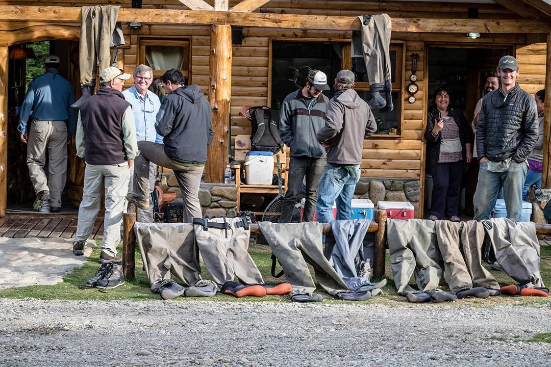 Guests gathered outside a rustic lodge, with waders and fishing gear laid out on the ground.