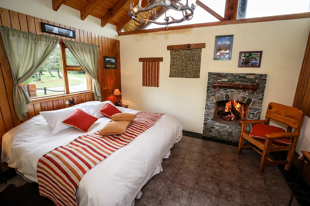 Cozy double room with a large bed, wood-paneled walls, stone fireplace, and a window overlooking greenery.