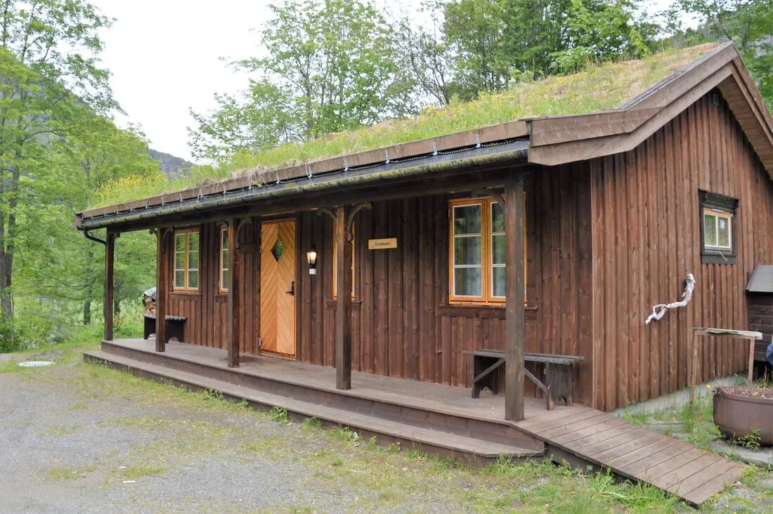 Small, rustic wooden cabin with a grass roof and external stairs leading up a grassy bank.