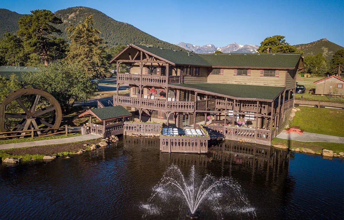 Aerial view of the lodge building on a pond with a water fountain, surrounded by mountains.