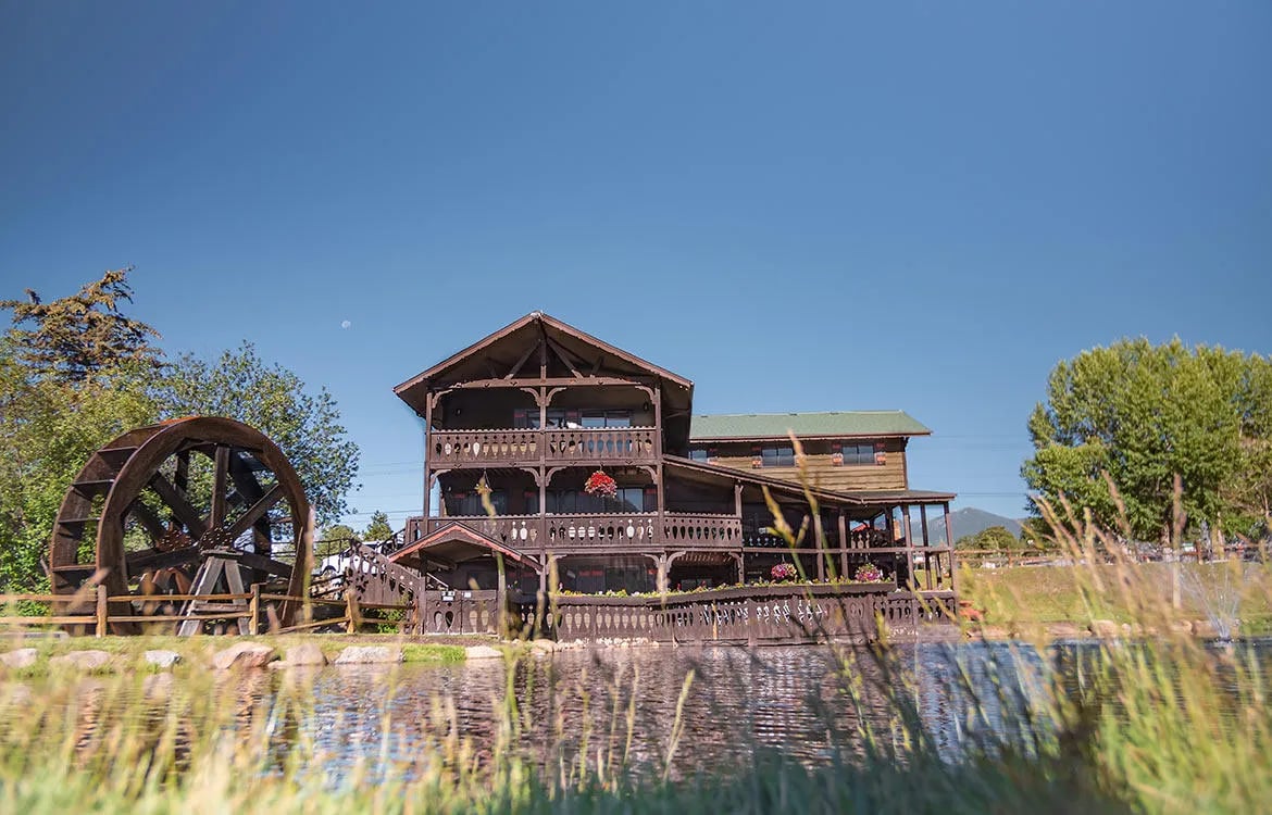 Large multi-story lodge building with a water wheel on a pond, seen from a grassy foreground.