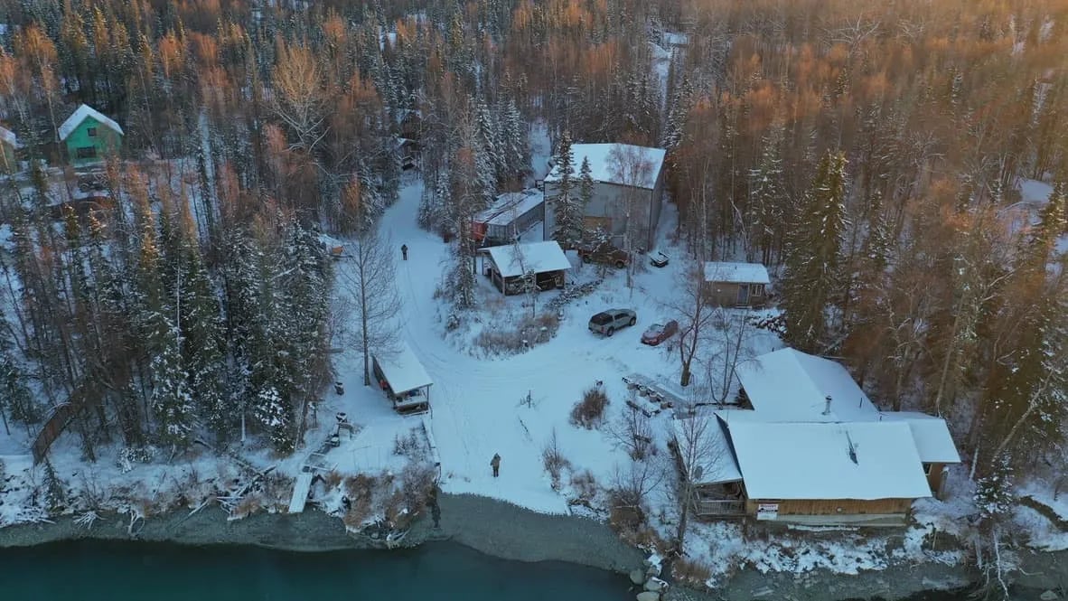 Aerial view of snow-covered lodge buildings nestled in a forest beside a river
