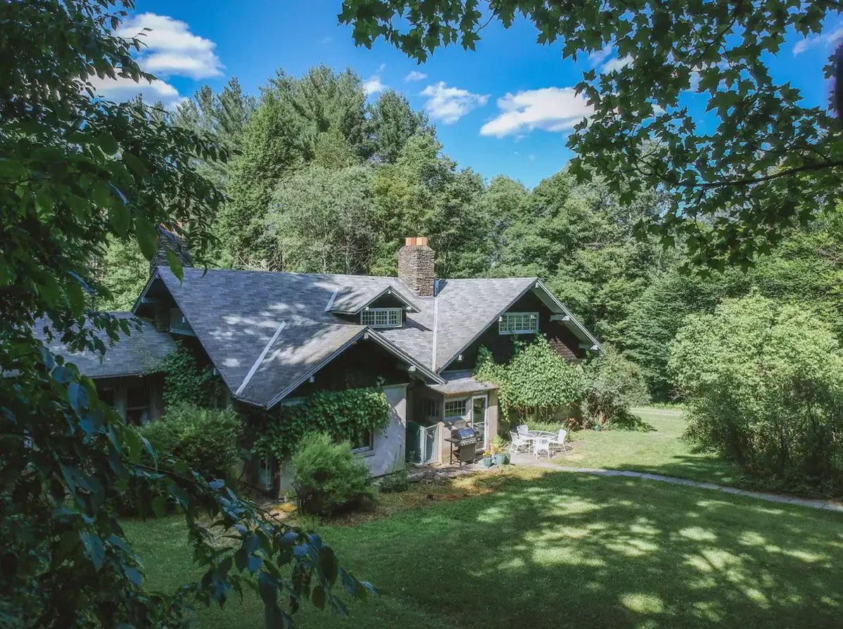 A house with a dark roof surrounded by trees and greenery on a sunny day, with patio furniture and a barbecue grill visible outside.