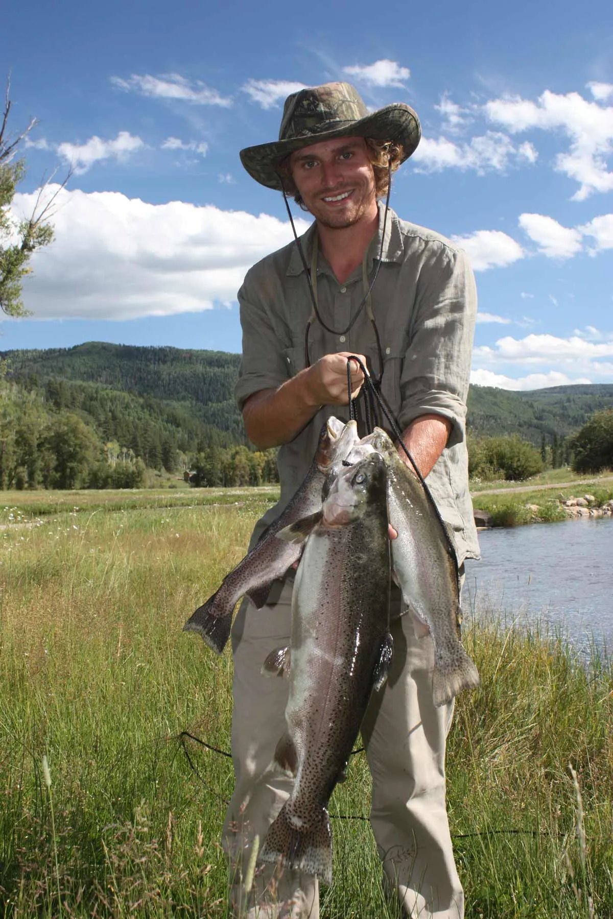 Man in a hat holding a stringer of three large fish by a grassy stream