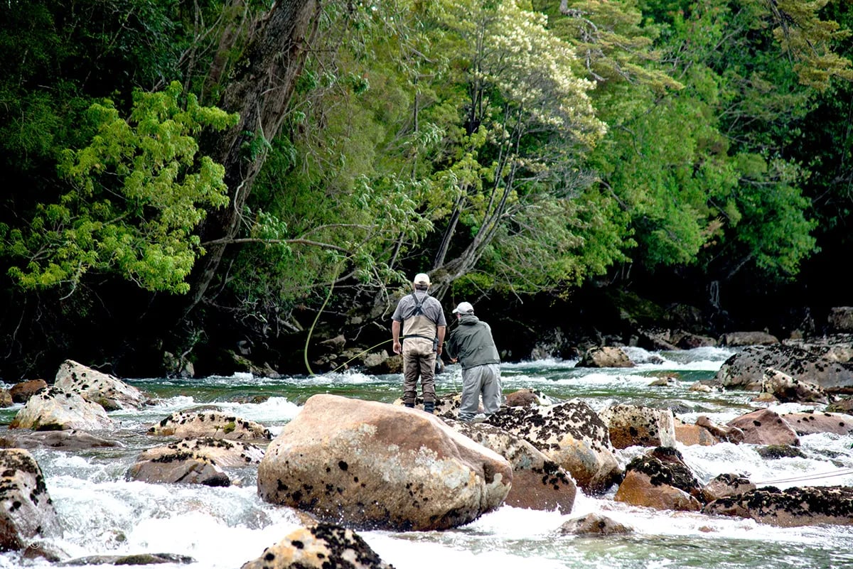 Two fly fishermen wading in a rocky river with lush green trees on the banks
