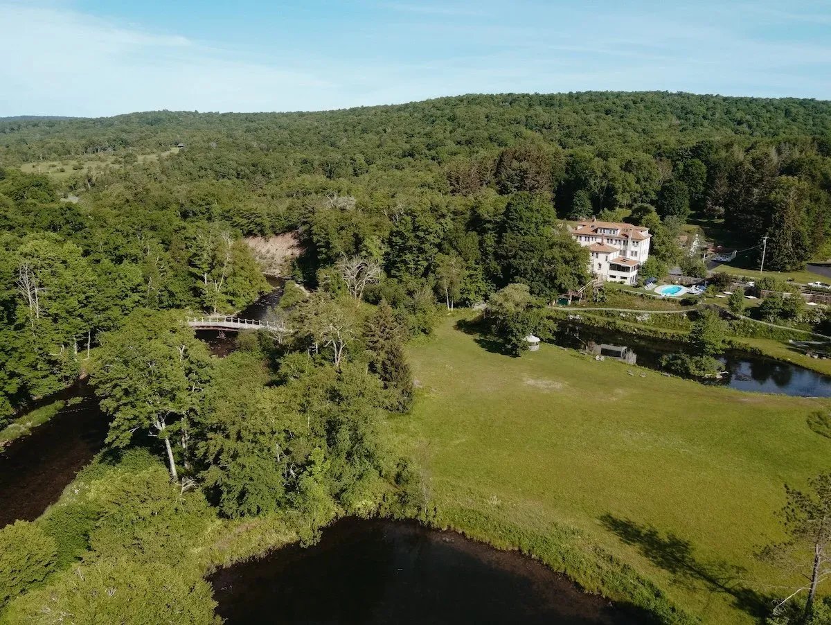 Aerial view of a green landscape with a river, a bridge, trees, a large house with a pool, and rolling hills in the background.