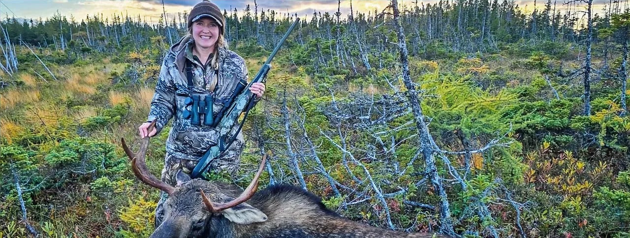 Hunter in camouflage standing over a harvested moose in a brushy field.