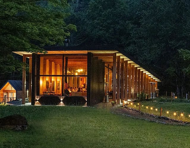A-frame cabin interior with a bed facing a large triangular window overlooking a winter forest.