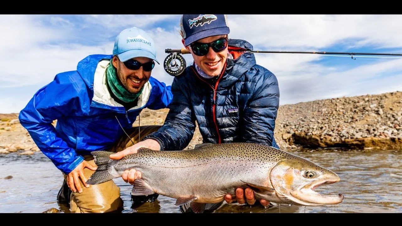 Two smiling anglers in waders holding a large rainbow trout in a shallow river