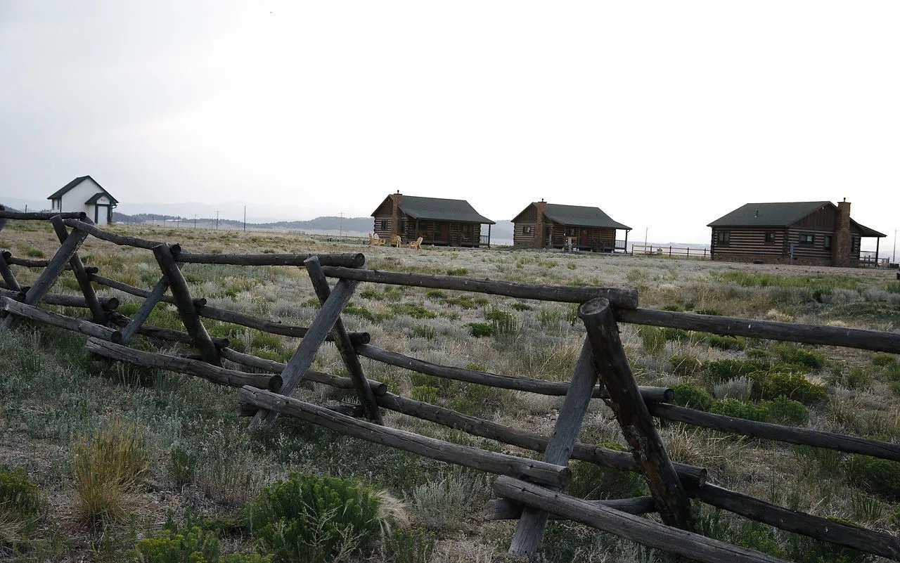 Several rustic cabins scattered across a grassy field with a wooden fence in the foreground.