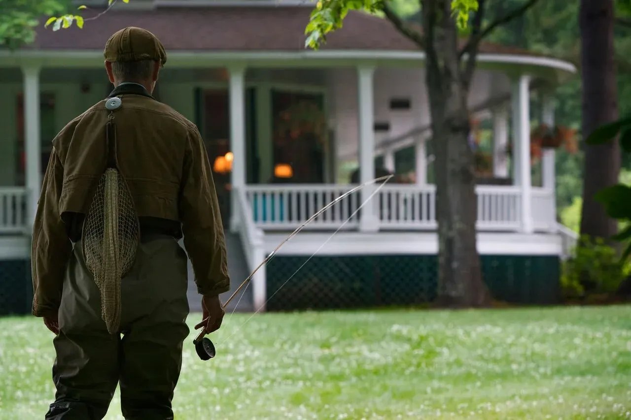 An angler in fishing gear standing on a lawn, holding a fishing rod, with the Beaverkill Valley Inn and trees in the background.