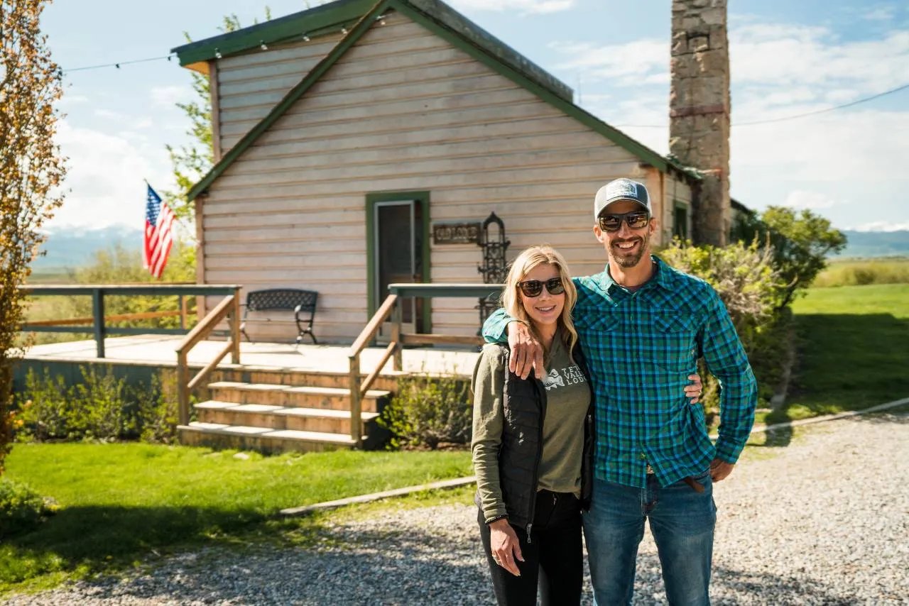 Couple smiling in front of a rustic lodge building with an American flag