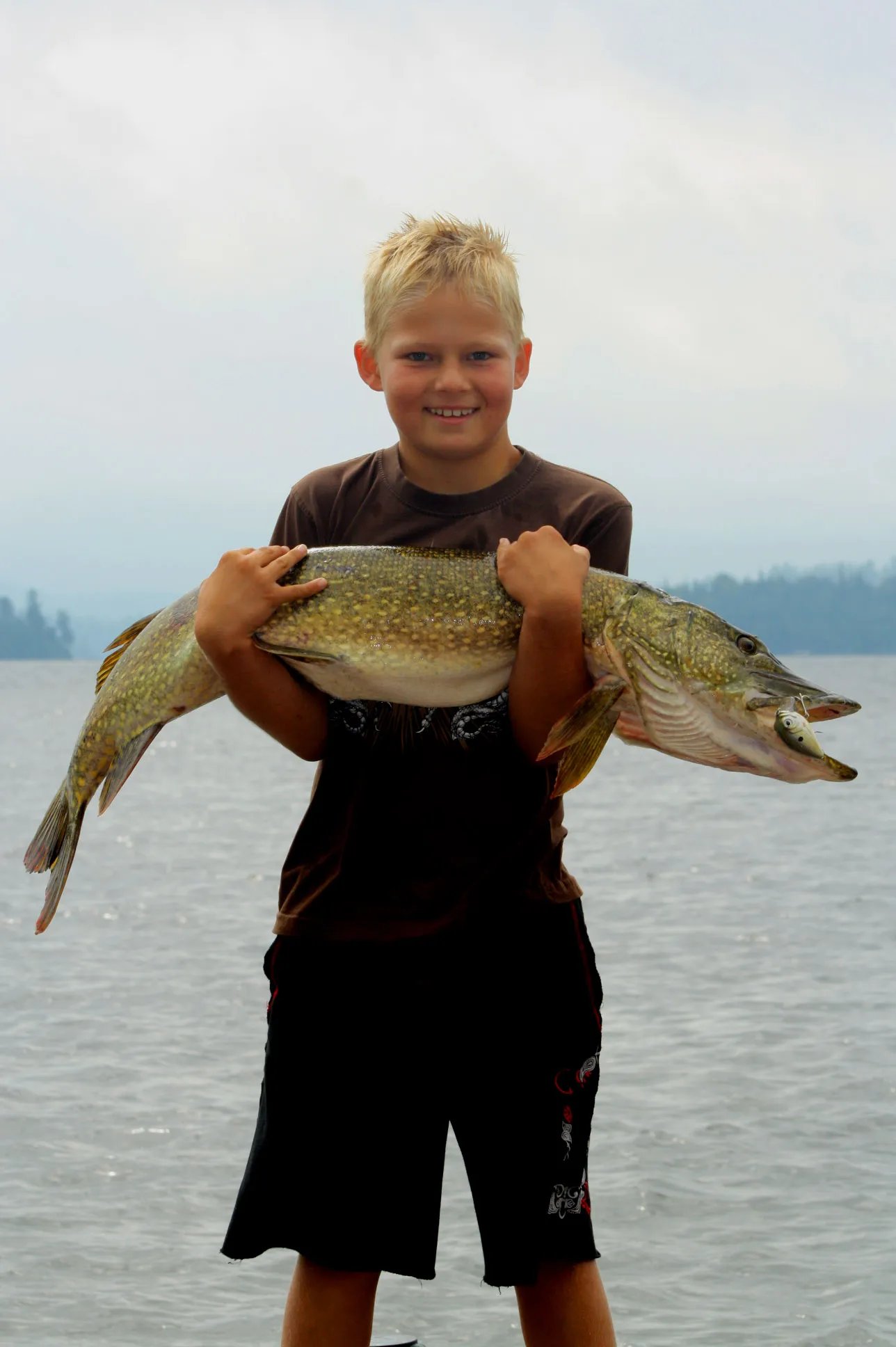 Smiling boy holding a large northern pike against a lake background
