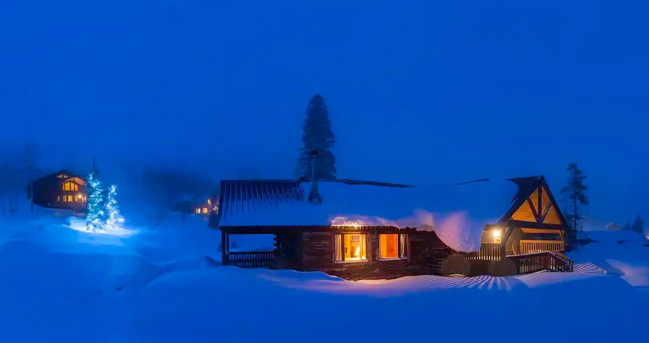 Snow-covered log cabin with warm lights glowing at dusk