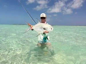 Smiling woman holding a bonefish in clear shallow water, wearing a hat and sunglasses