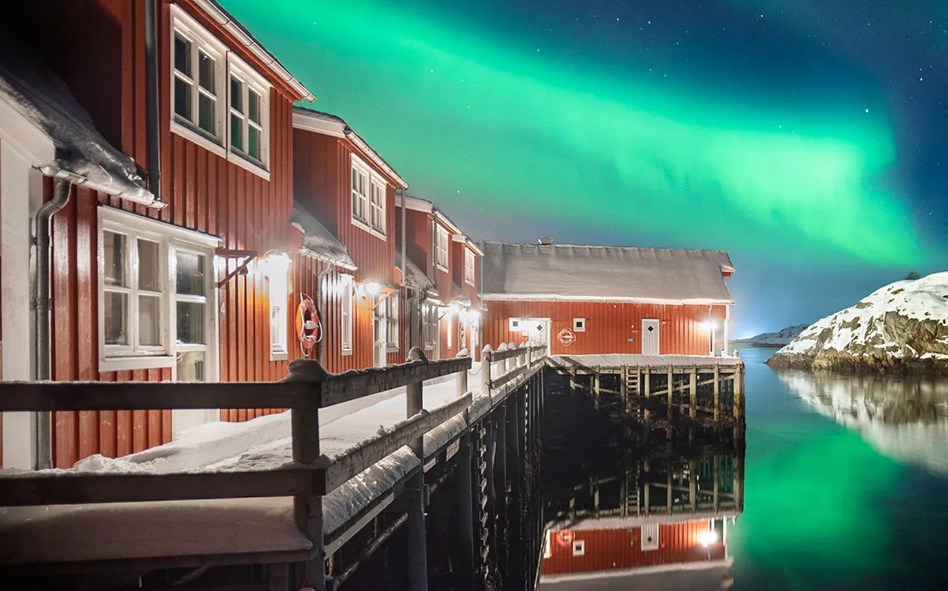 Red cabins on stilts over calm water, reflecting green Northern Lights in the night sky