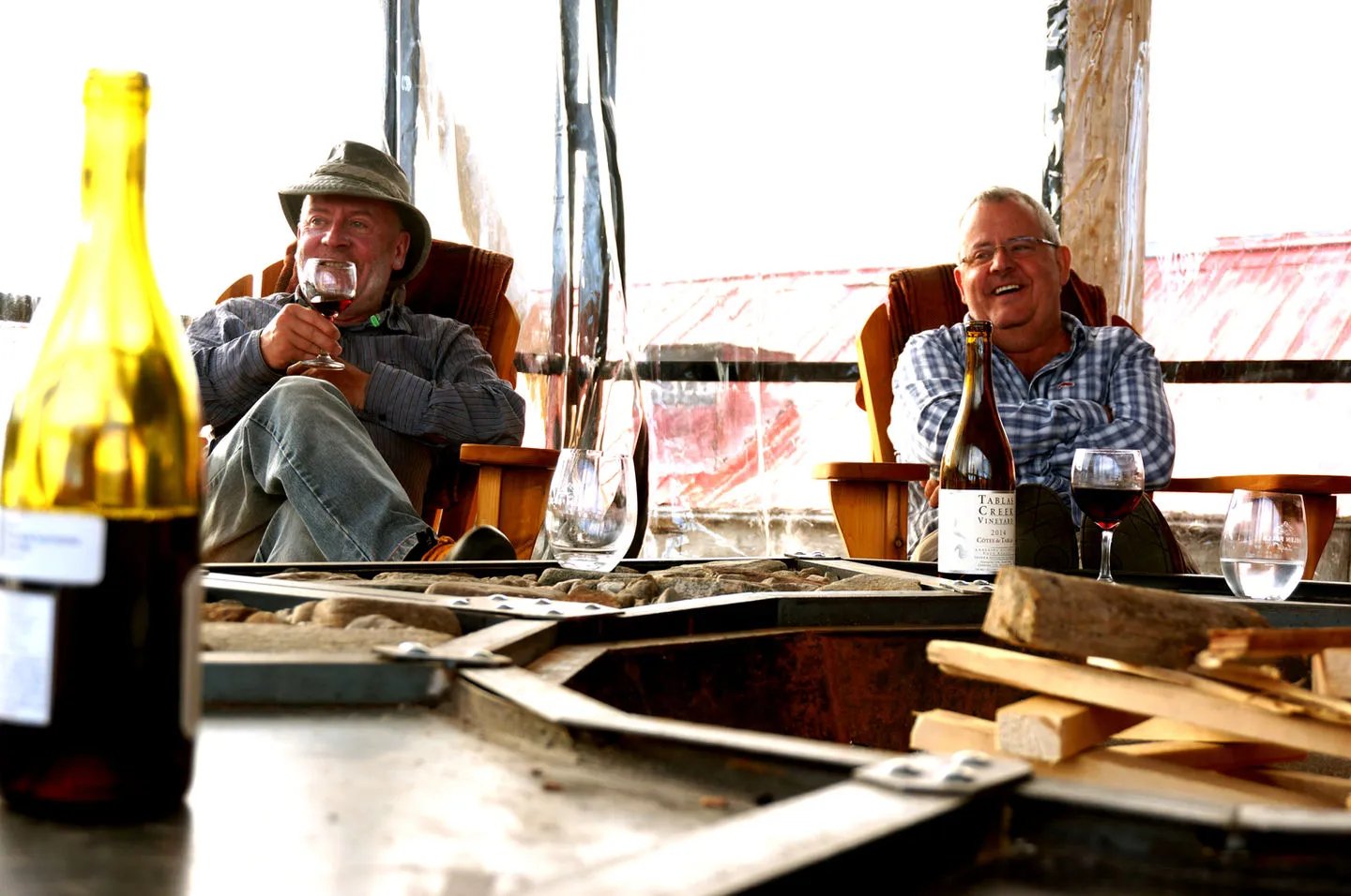 Two men relaxing by an indoor fire pit with wine bottles, enjoying conversation