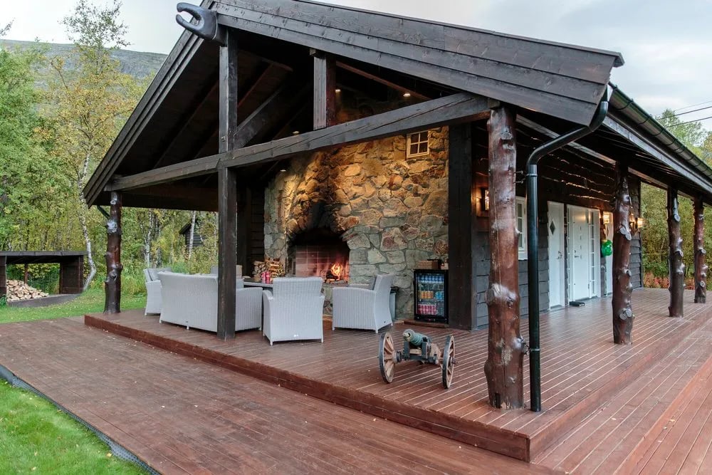 Dark wood cabin with a covered porch and deck surrounded by green foliage.