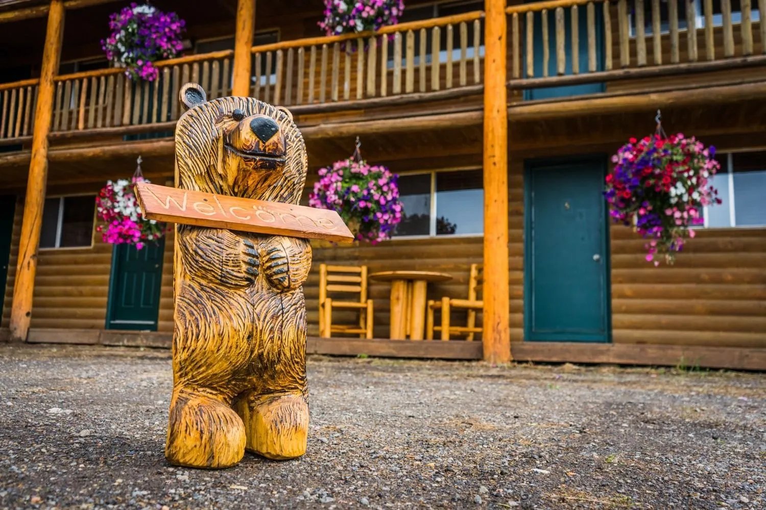 Carved wooden bear statue holding a sign outside a rustic log lodge with hanging flower baskets