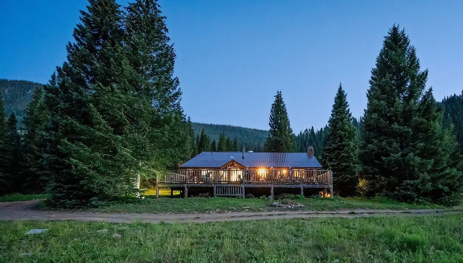 Main lodge building glowing with warm light at dusk, surrounded by tall pine trees.