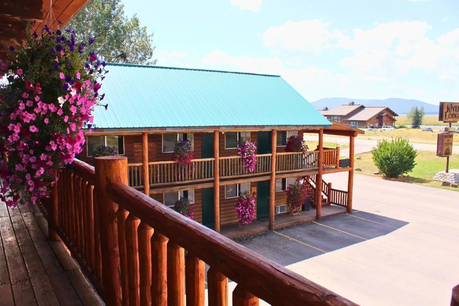 Two-story log motel building with a teal roof and balconies adorned with pink hanging flowers
