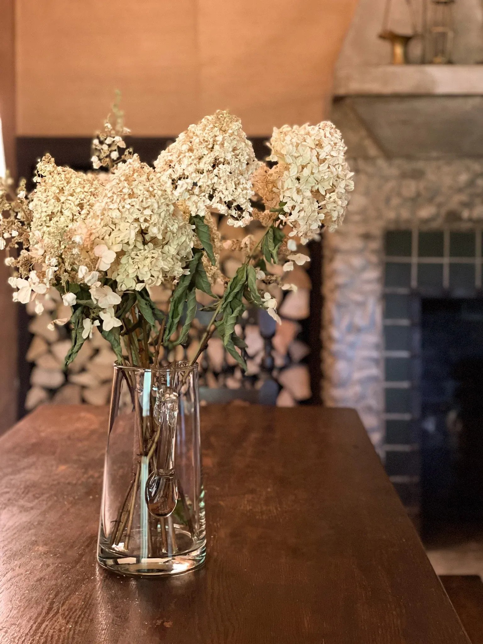 A clear glass vase with dried white hydrangea flowers on a wooden table in a cozy room with a stone fireplace and firewood stacked in the background.