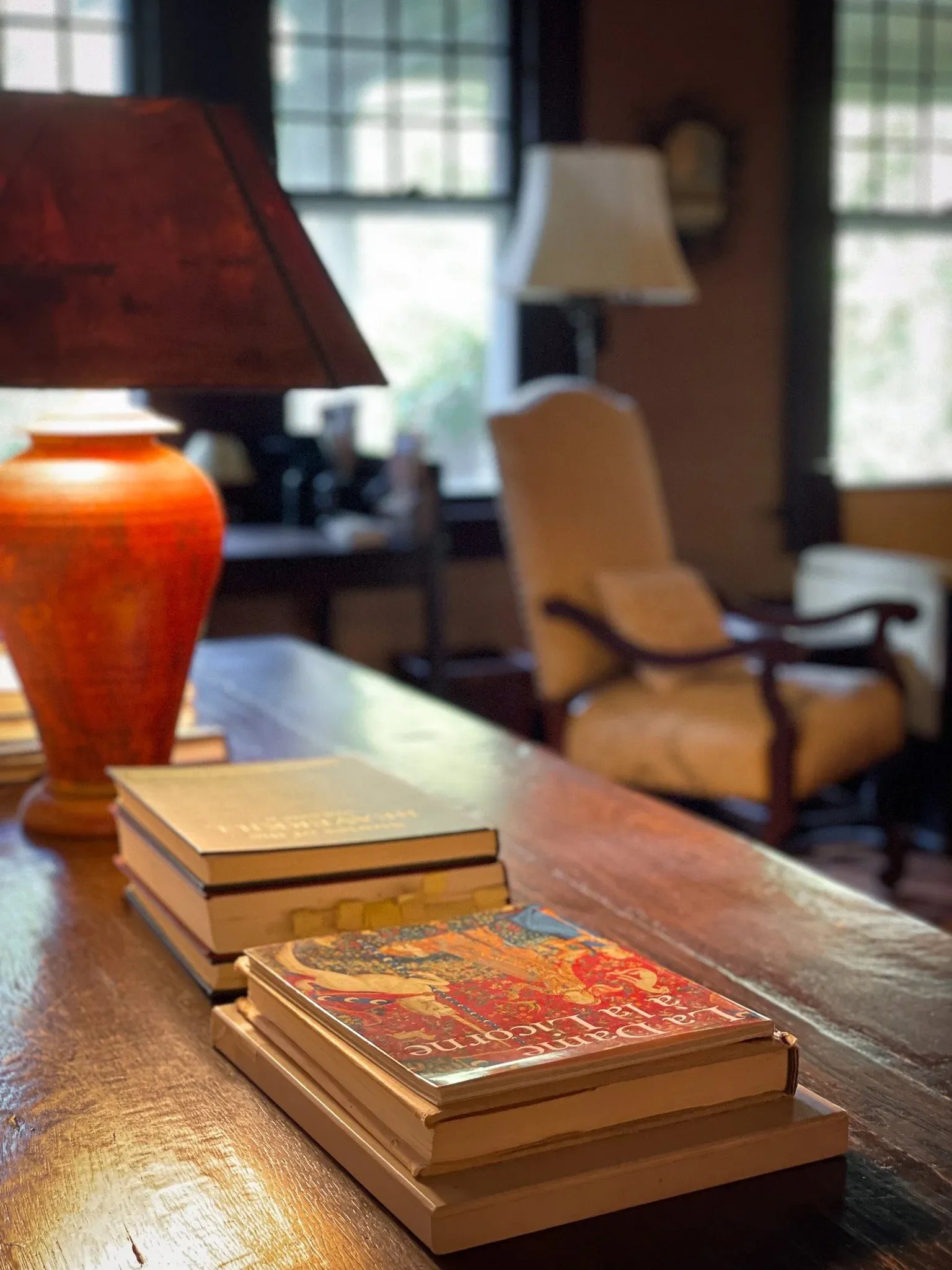 Stack of four hardcover books on a wooden table in a cozy, warmly lit room with armchairs and large windows in the background.