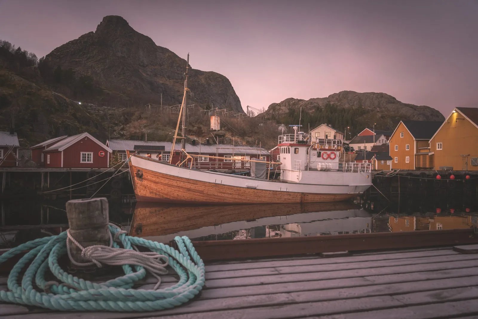 Traditional fishing boat docked in Nusfjord harbor at dusk with mountains in the background