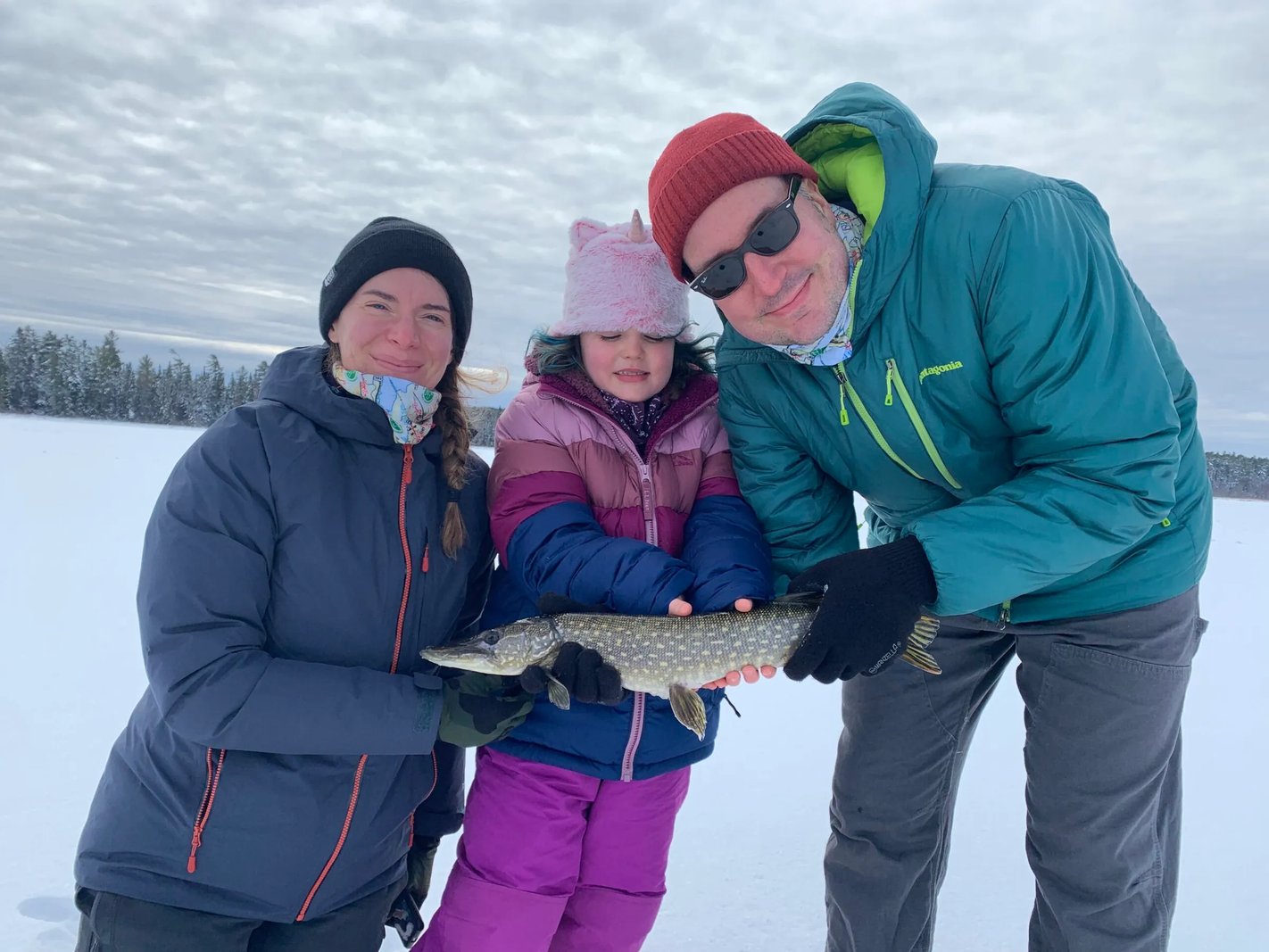 Family of three holding a Northern Pike on a snowy frozen lake