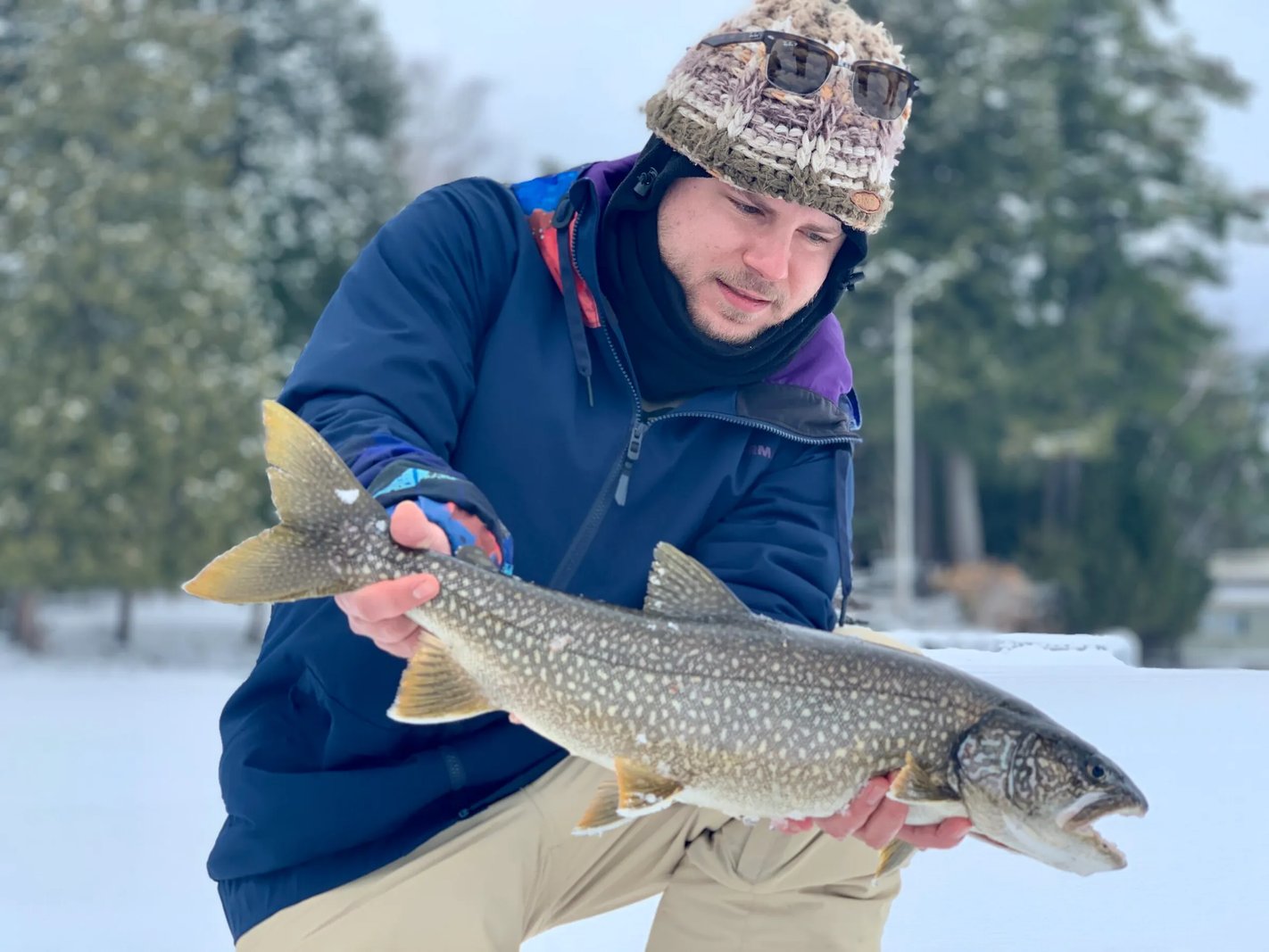 Man in a winter hat holding a large Lake Trout on a snowy lake