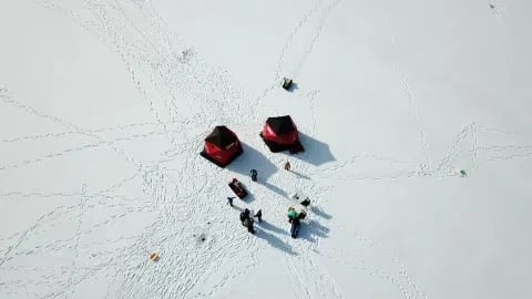Angler sitting by a red ice fishing tent and gear on a wide, snowy frozen lake