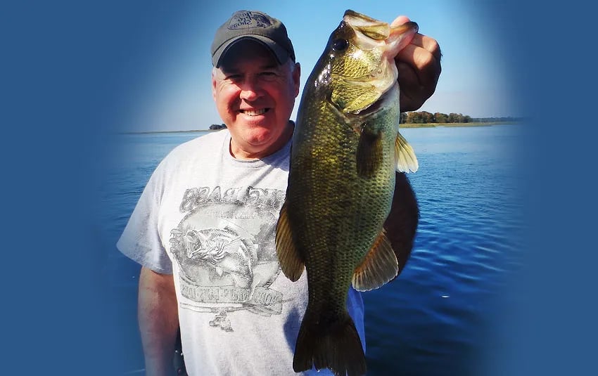 Man in a cap smiling, holding a largemouth bass over blue water.